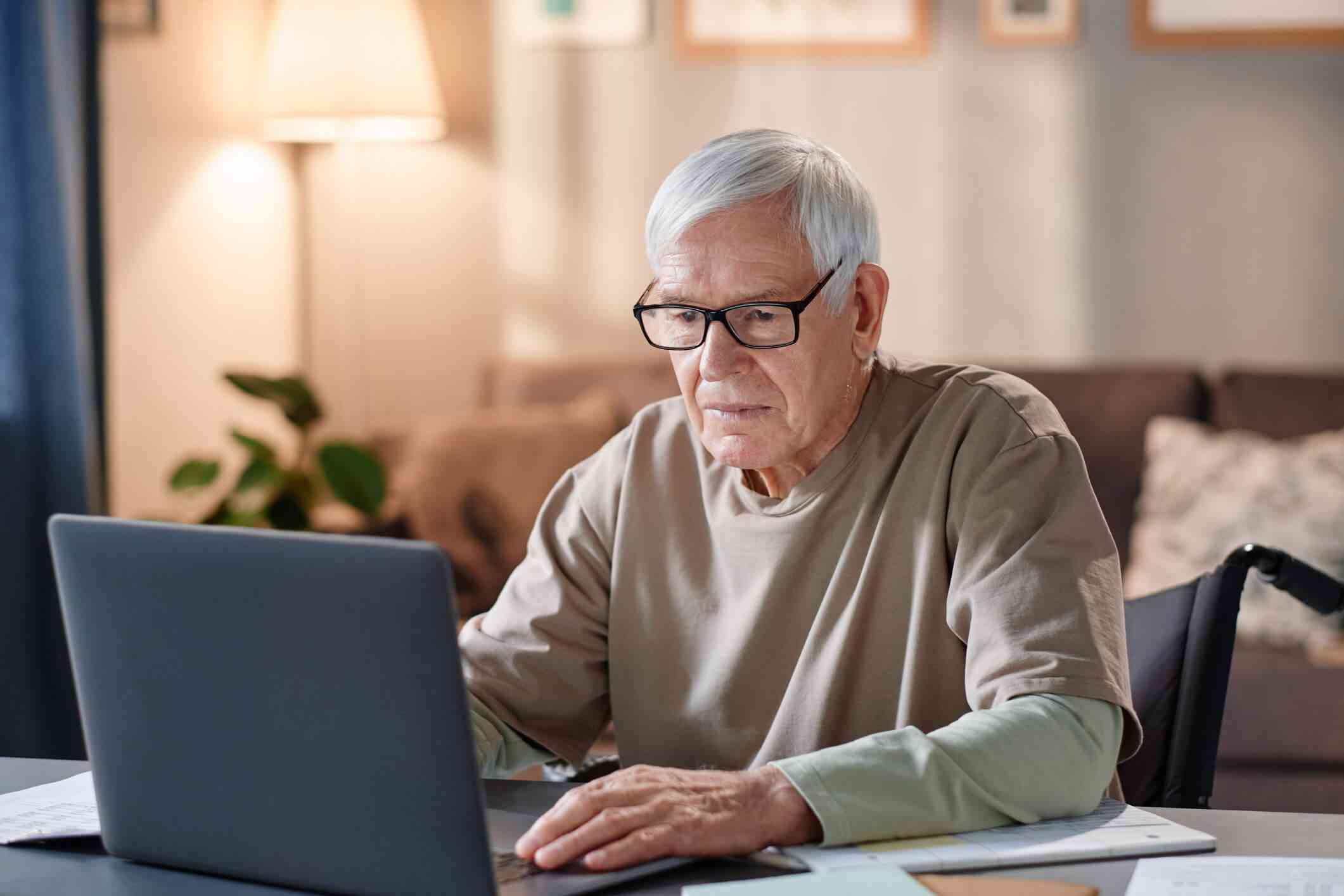 An elderly man sits in his wheelchair at a table and types on the open laptop infront of him.