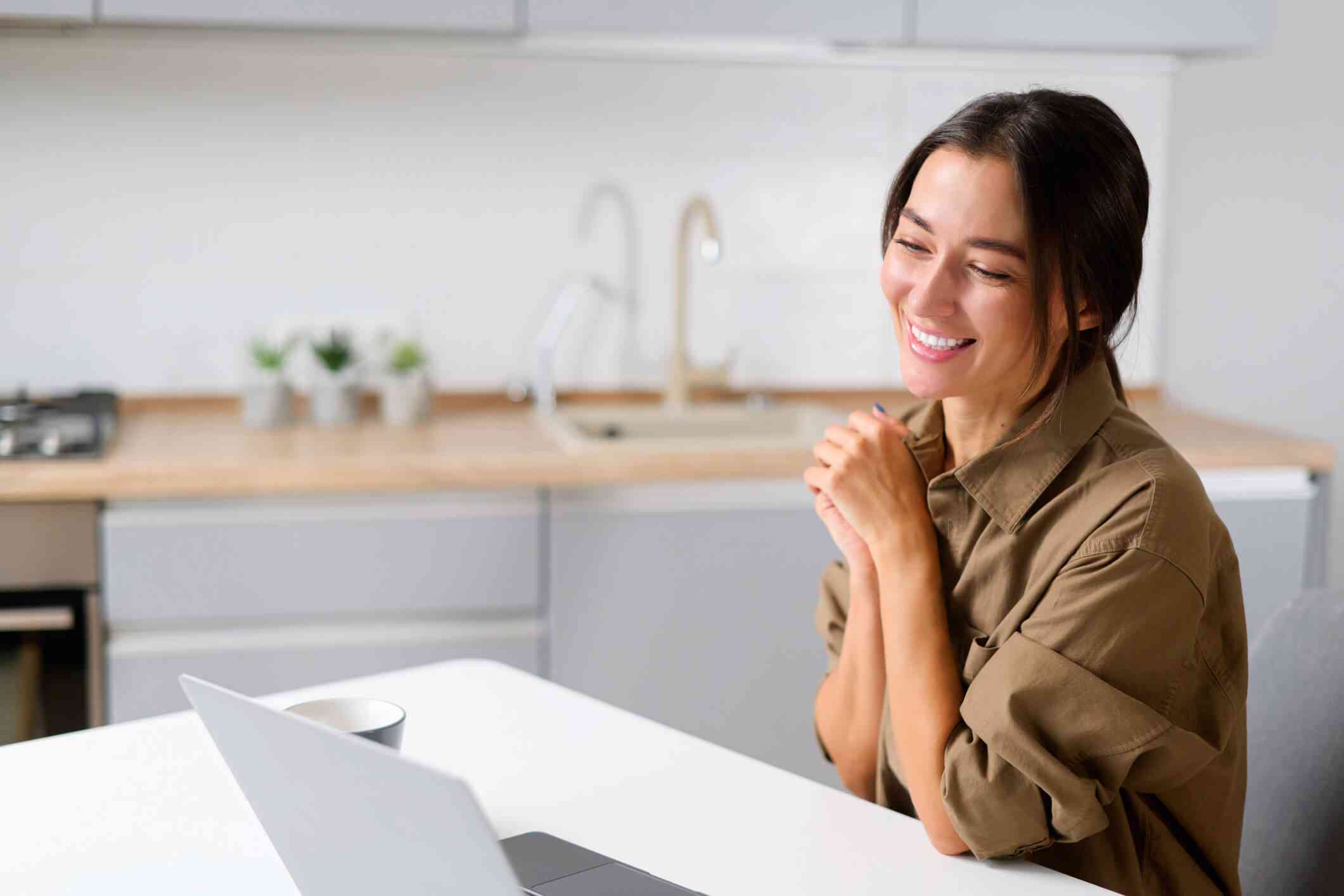 Woman in a brown button up shirt sits at her kitchen counter and smiles while she talks to a therapist on her computer