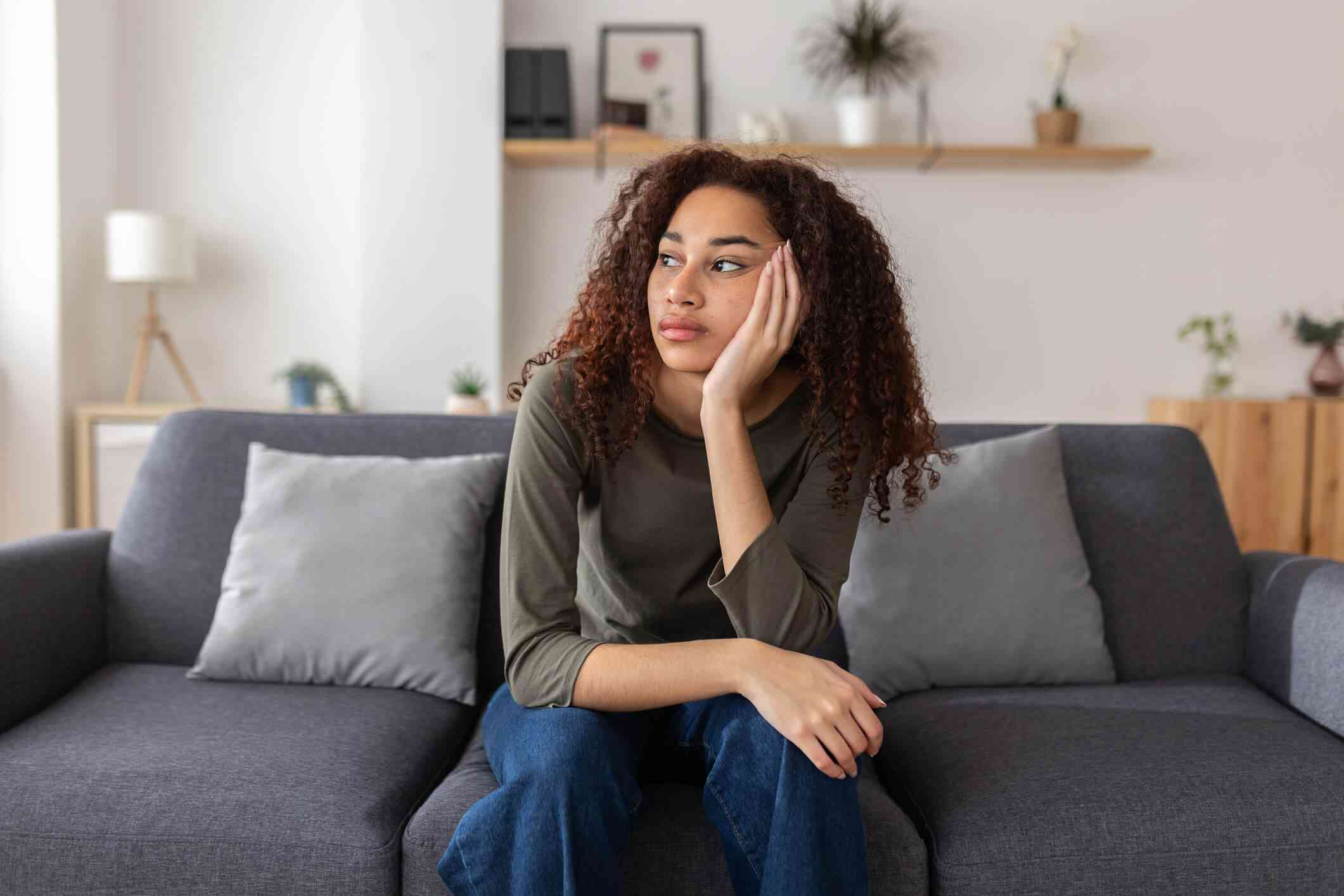 A woman in a grey shirt sits sadly on her couch with her head in her hand while gazing off sadly.
