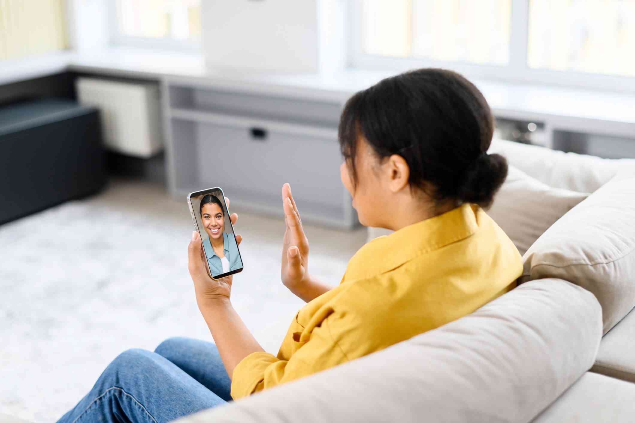 A woman in a yellow shirt sits on her couch and waves at her therpaist on the phone in her other hand during a virtual therpay session.
