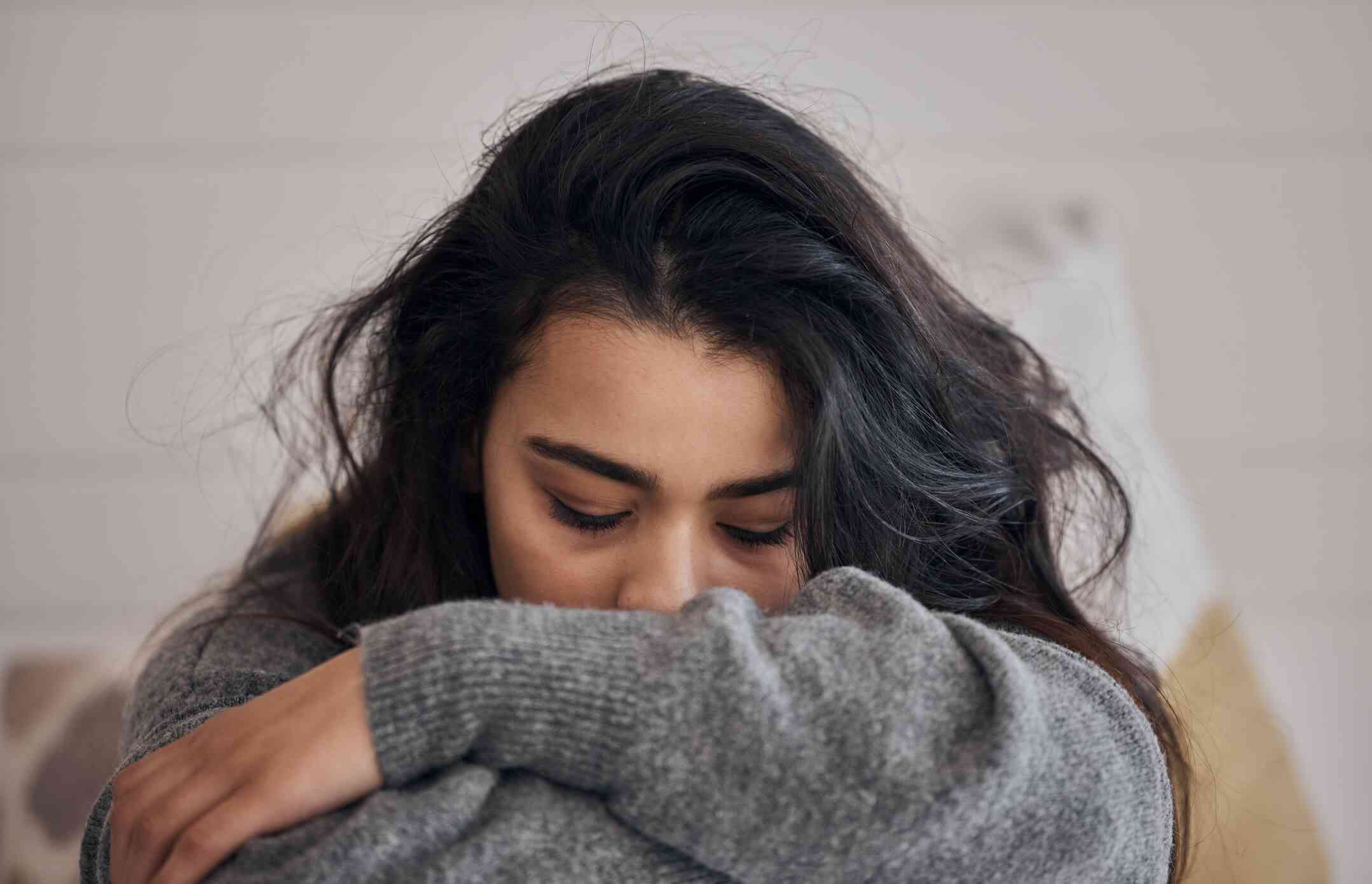 A woman in a grey sweater sadly closes her eyes and hides her face in her arms.